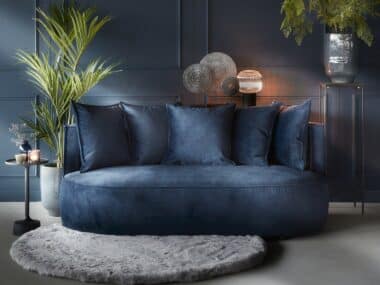 Velvet donker blauwe tweezitsbank met ronde vormen en bijhorende rugkussens. Gestyled met een rond vloerkleed en planten.