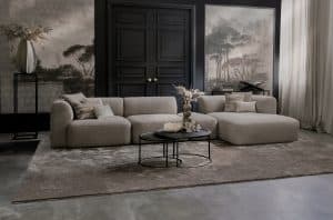 Elementenbank Emanuelle met een longchair en een hocker tussen de elementen in. Uitgevoerd in een lichte beige stof. Gecombineerd met een groot vloerkleed en naturel decoratie.