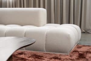Lichte hoekbank Oriane met warme sierkussens op een bordeaux vloerkleed in een moderne woonkamer.