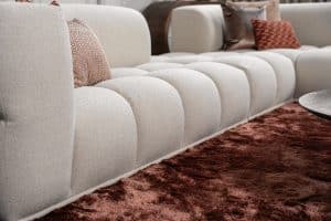 Lichte hoekbank Oriane met warme sierkussens op een bordeaux vloerkleed in een moderne woonkamer.