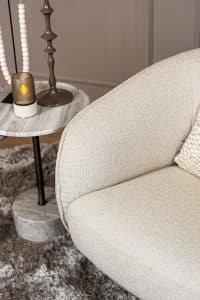 Lio draaifauteuil van Gusj in lichte bouclé stof met anodic brown draaionderstel op een rond vloerkleed in een warm interieur.